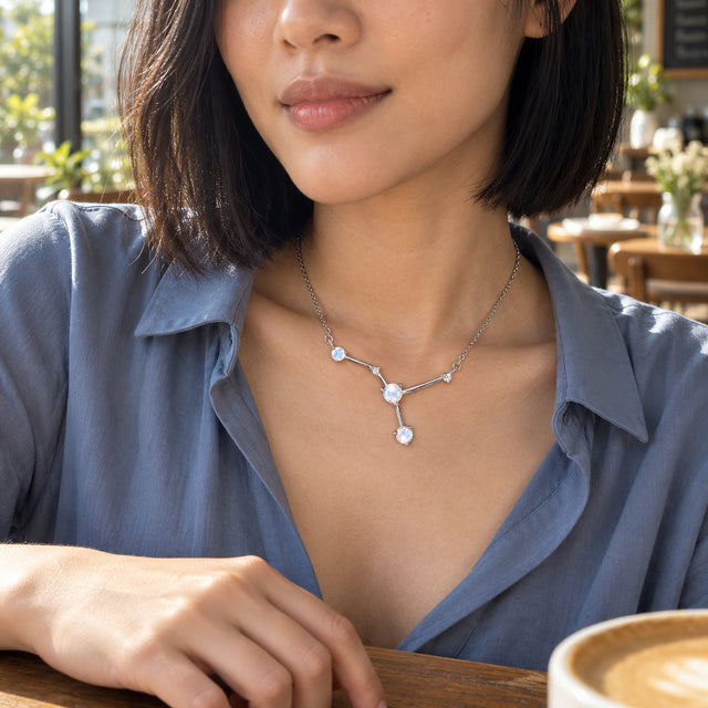 A woman in a blue shirt wears a silver branching moonstone necklace at a cafe, with a ceramic cup on the wooden table.