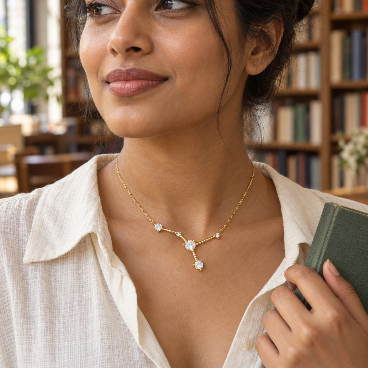 A woman in a white linen shirt wears a yellow gold branching moonstone necklace while holding a green book beside bookshelves.