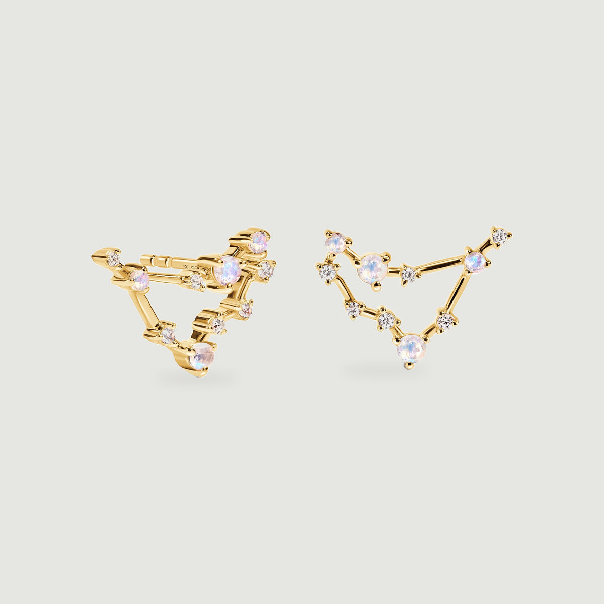 Two yellow gold stud earrings show open angular outlines linked by moonstones and clear accents above soft shadows.
