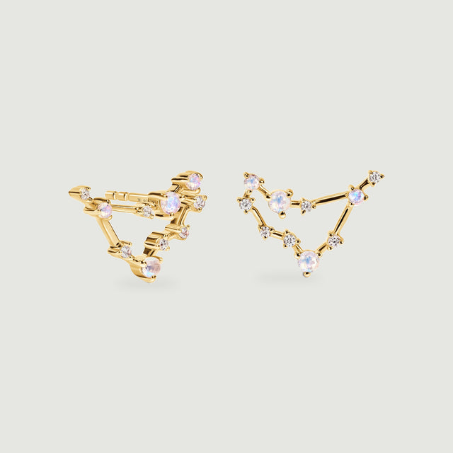 Two yellow gold stud earrings show open angular outlines linked by moonstones and clear accents above soft shadows.