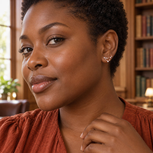 A woman with short coiled hair wears a rose gold constellation stud beside wooden bookshelves.