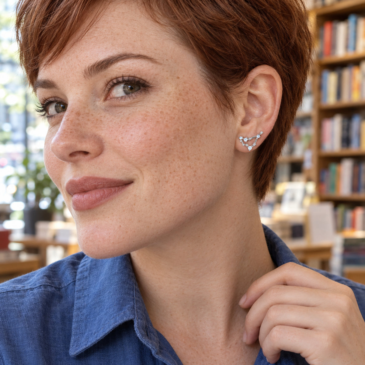 A freckled woman with short auburn hair wears a silver constellation earring in a bookstore.