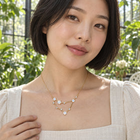 A woman in a cream linen blouse wears a yellow gold geometric necklace among sunlit green plants.