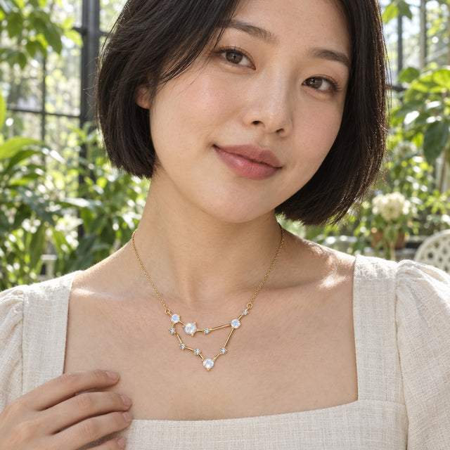 A woman in a cream linen blouse wears a yellow gold geometric necklace among sunlit green plants.