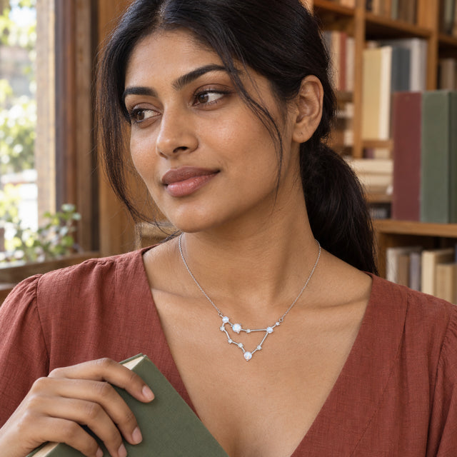 A woman in a terracotta blouse wears a silver necklace while holding a green book beside wooden shelves.