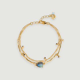 A yellow gold bracelet with two linked strands, dark bead accents and a blue-grey labradorite centre rests on a pale grey background.