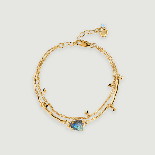 A yellow gold bracelet with two linked strands, dark bead accents and a blue-grey labradorite centre rests on a pale grey background.