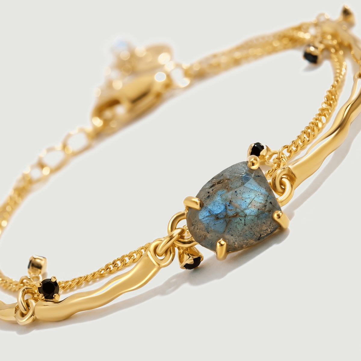 An angled view shows the blue-grey stone held by gold prongs beside a fine chain and polished curved links.