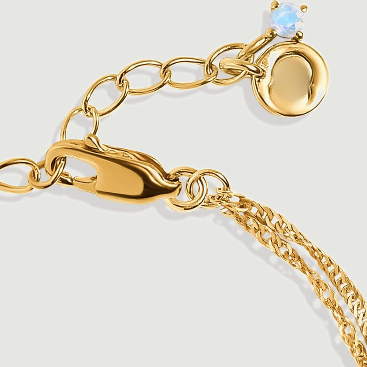 A yellow gold lobster clasp connects oval extension links ending in a small pale blue stone and a polished round charm.