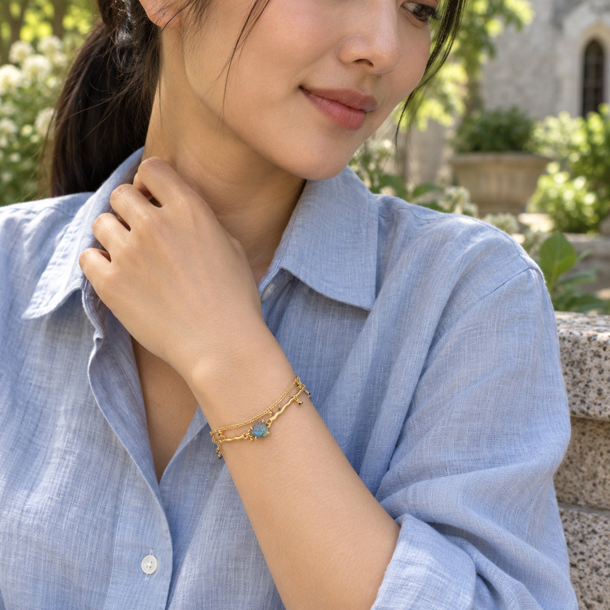 A woman in a pale blue shirt wears the yellow gold labradorite bracelet on her raised wrist in a leafy courtyard.
