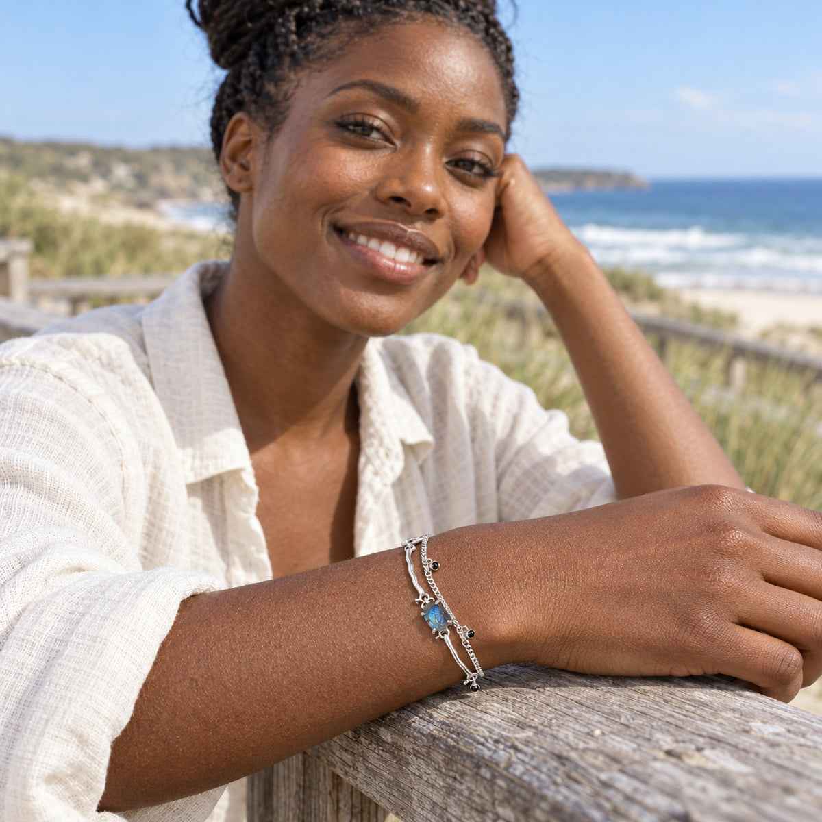 A woman in a cream linen shirt rests her wrist with the silver labradorite bracelet on a weathered wooden rail beside the sea.