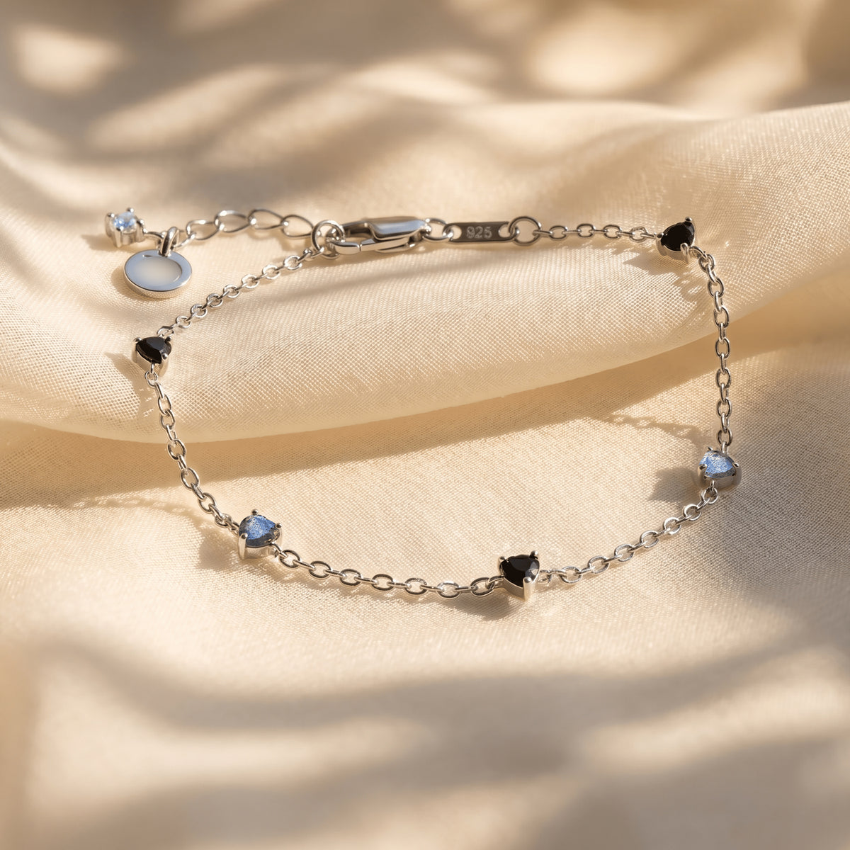The silver bracelet rests on folded cream fabric with its black and blue gray heart shaped stones visible.