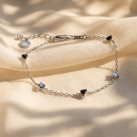 The silver bracelet rests on folded cream fabric with its black and blue gray heart shaped stones visible.