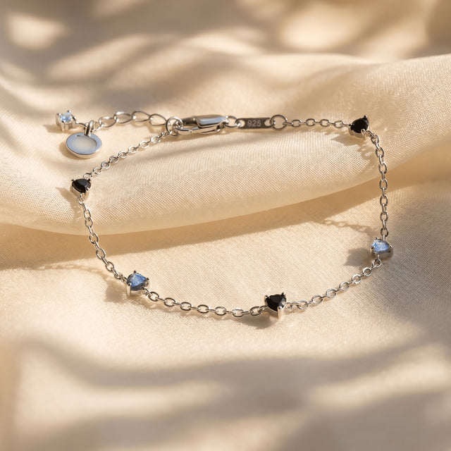 The silver bracelet rests on folded cream fabric with its black and blue gray heart shaped stones visible.