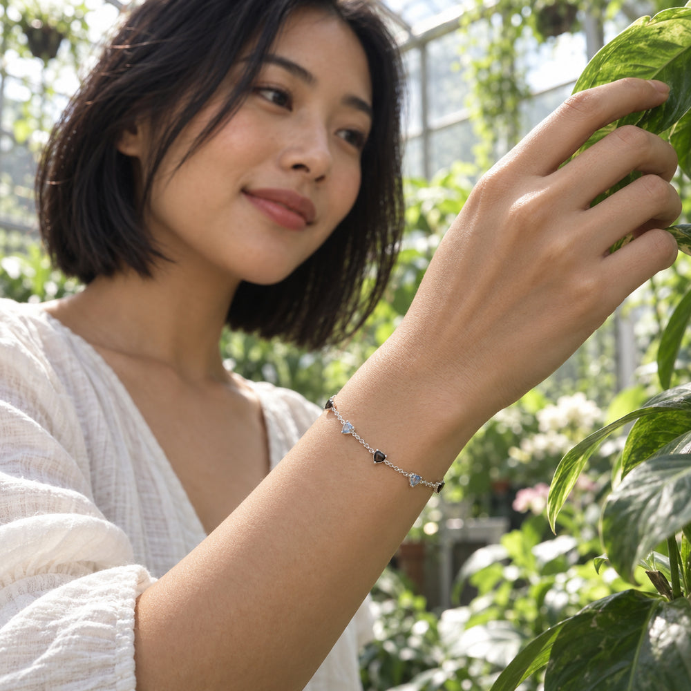 A silver bracelet with black and blue gray heart shaped stones is worn on a wrist among greenhouse plants.