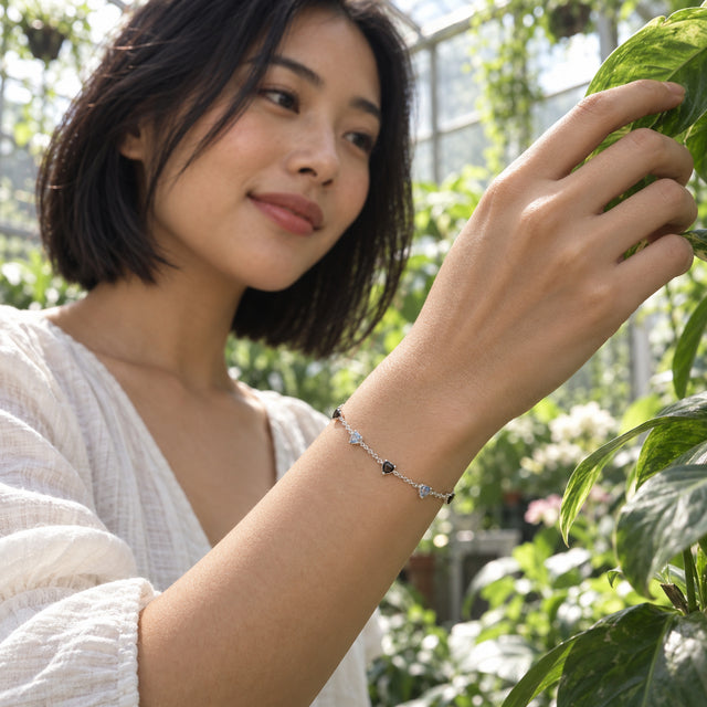 A silver bracelet with black and blue gray heart shaped stones is worn on a wrist among greenhouse plants.