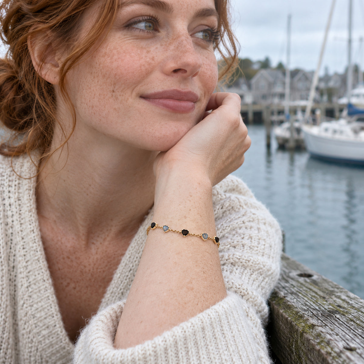 A yellow gold bracelet with alternating black and blue gray heart shaped stones circles a freckled wrist beside a harbor railing.