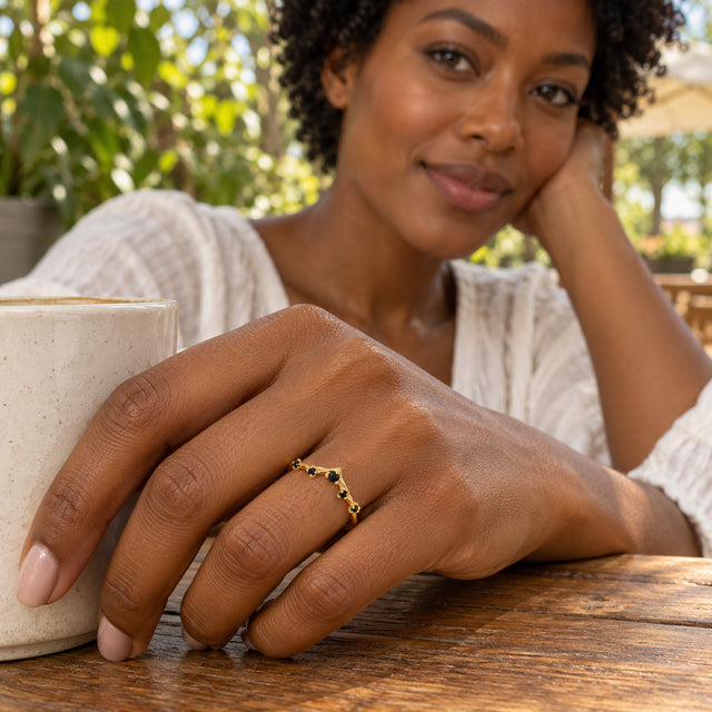 A woman with short curls wears a yellow gold ring with five black stones beside a ceramic cup at an outdoor cafe.
