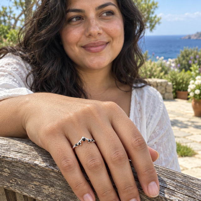 A silver ring with five black stones is worn on a woman's hand resting on a weathered wooden bench beside the sea.