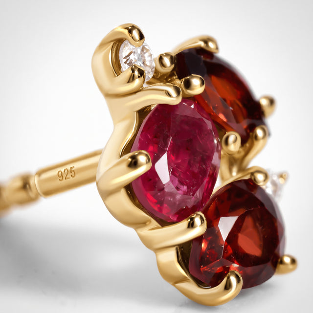 A close view of yellow gold prongs surrounds a pink stone, dark red facets and a clear accent above a post stamped 925.