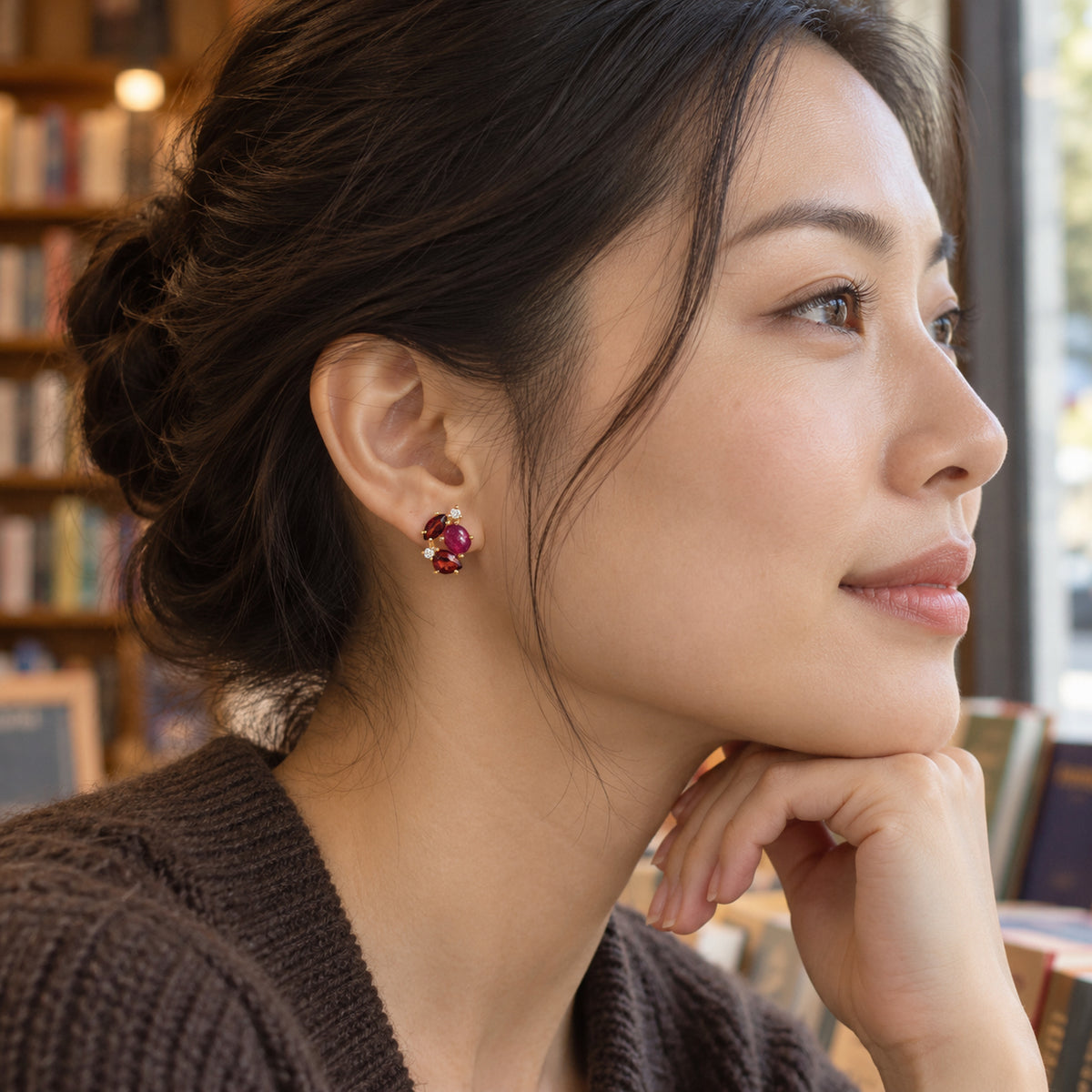 A woman with dark hair in a bun wears a yellow gold gemstone stud beside a bookstore window, with her hand resting under her chin.