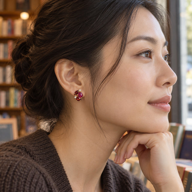 A woman with dark hair in a bun wears a yellow gold gemstone stud beside a bookstore window, with her hand resting under her chin.