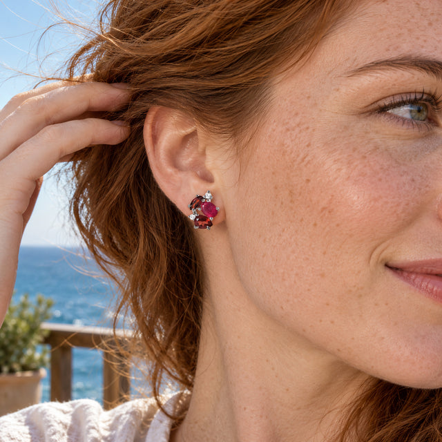 A woman with auburn hair lifts it away from a silver gemstone stud on a sunlit terrace overlooking the sea.