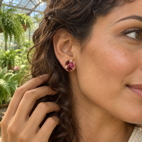 A woman with curly brown hair wears a rose gold gemstone stud while holding a curl in a leafy greenhouse.