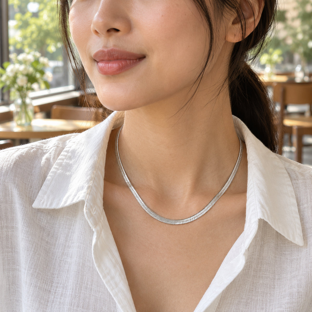 A woman wears a silver colored herringbone chain with an open white linen shirt beside a cafe window.