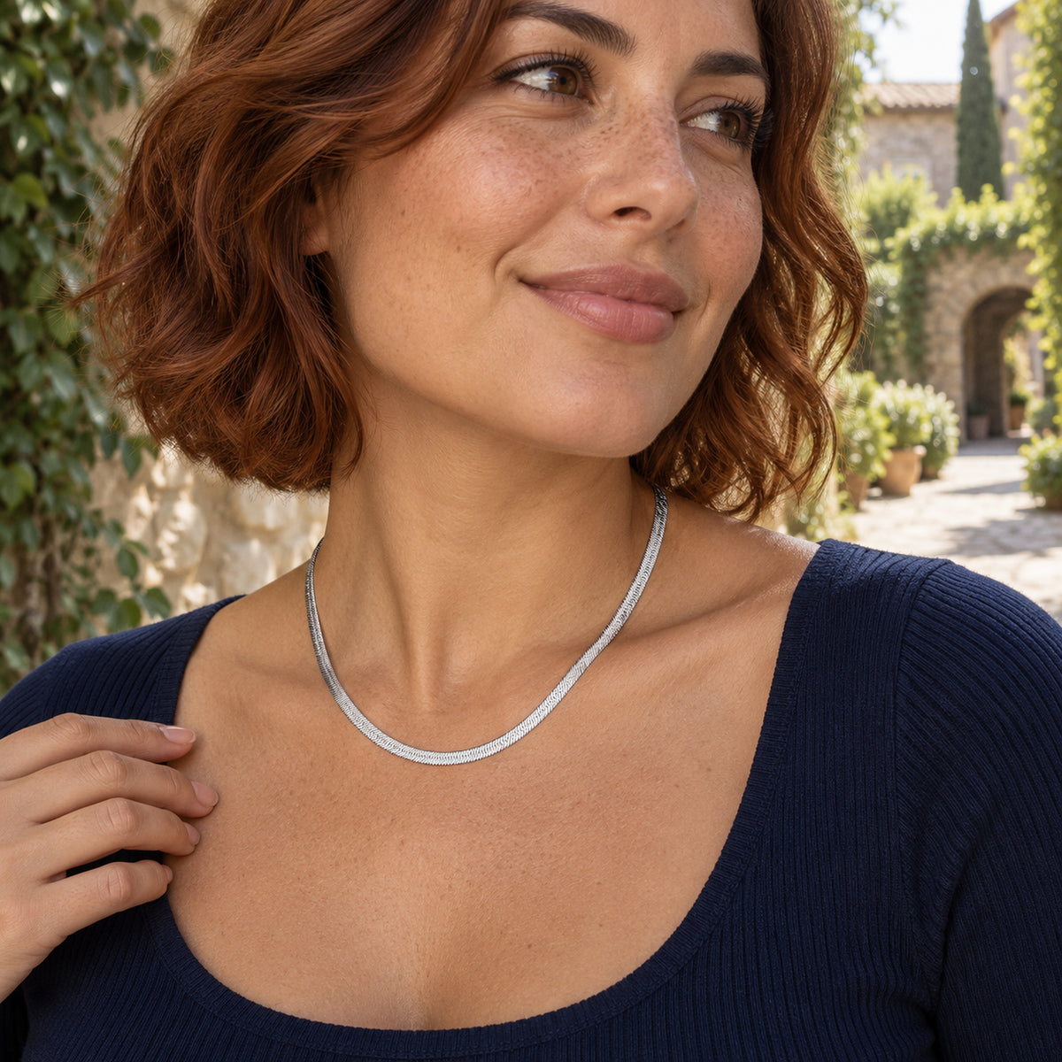 A woman in a navy top wears a silver colored flat chain in a sunlit stone courtyard.