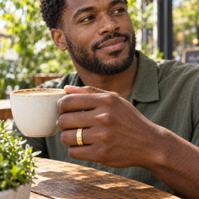A man wears a faceted yellow gold ring while holding a ceramic coffee cup at an outdoor cafe.