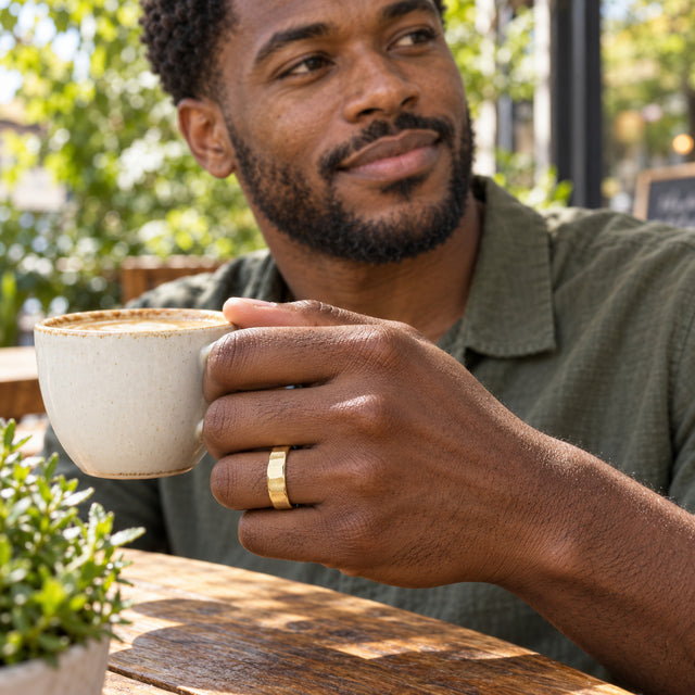 A man wears a faceted yellow gold ring while holding a ceramic coffee cup at an outdoor cafe.