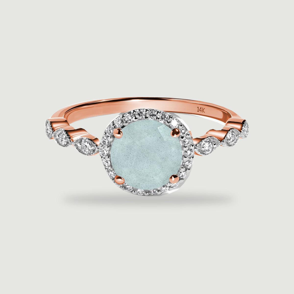 A rose gold ring with a pale blue oval aquamarine, a diamond halo, and curved diamond shoulders rests on a light gray surface.