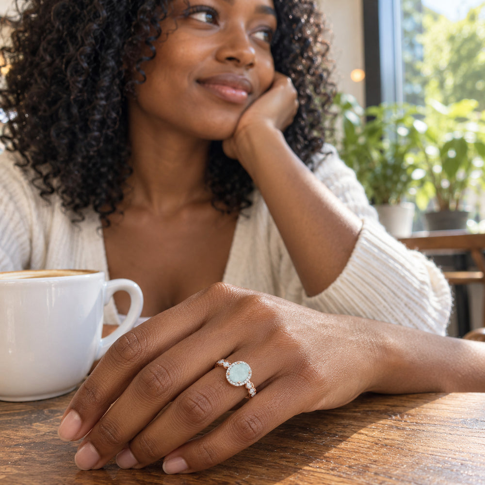 A woman with curly hair wears a rose gold aquamarine ring on her hand beside a white coffee cup at a wooden cafe table.