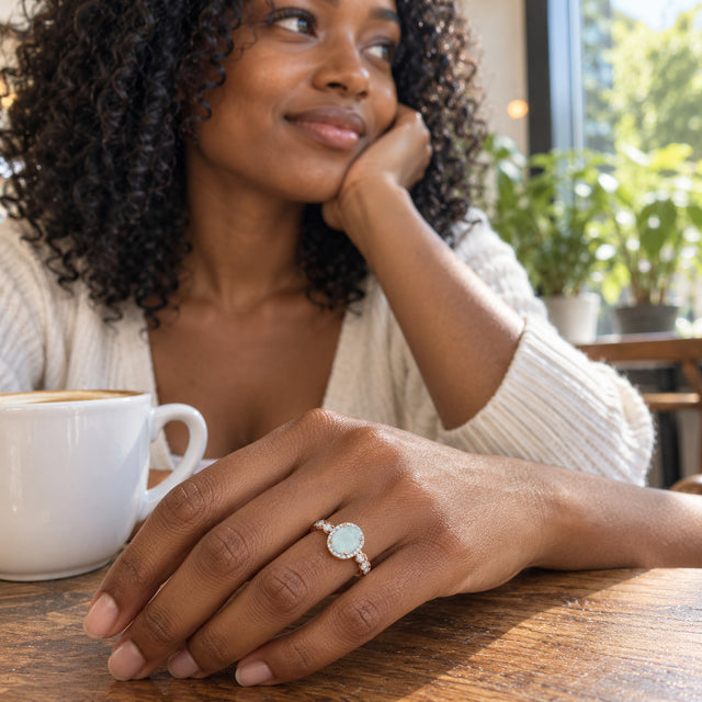 A woman with curly hair wears a rose gold aquamarine ring on her hand beside a white coffee cup at a wooden cafe table.
