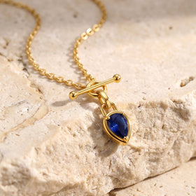 A gold-tone necklace lies diagonally on textured cream stone beside its blue pendant.
