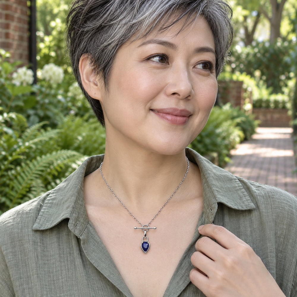 A woman in a sage linen blouse wears a silver-tone blue pendant necklace beside a leafy garden path.