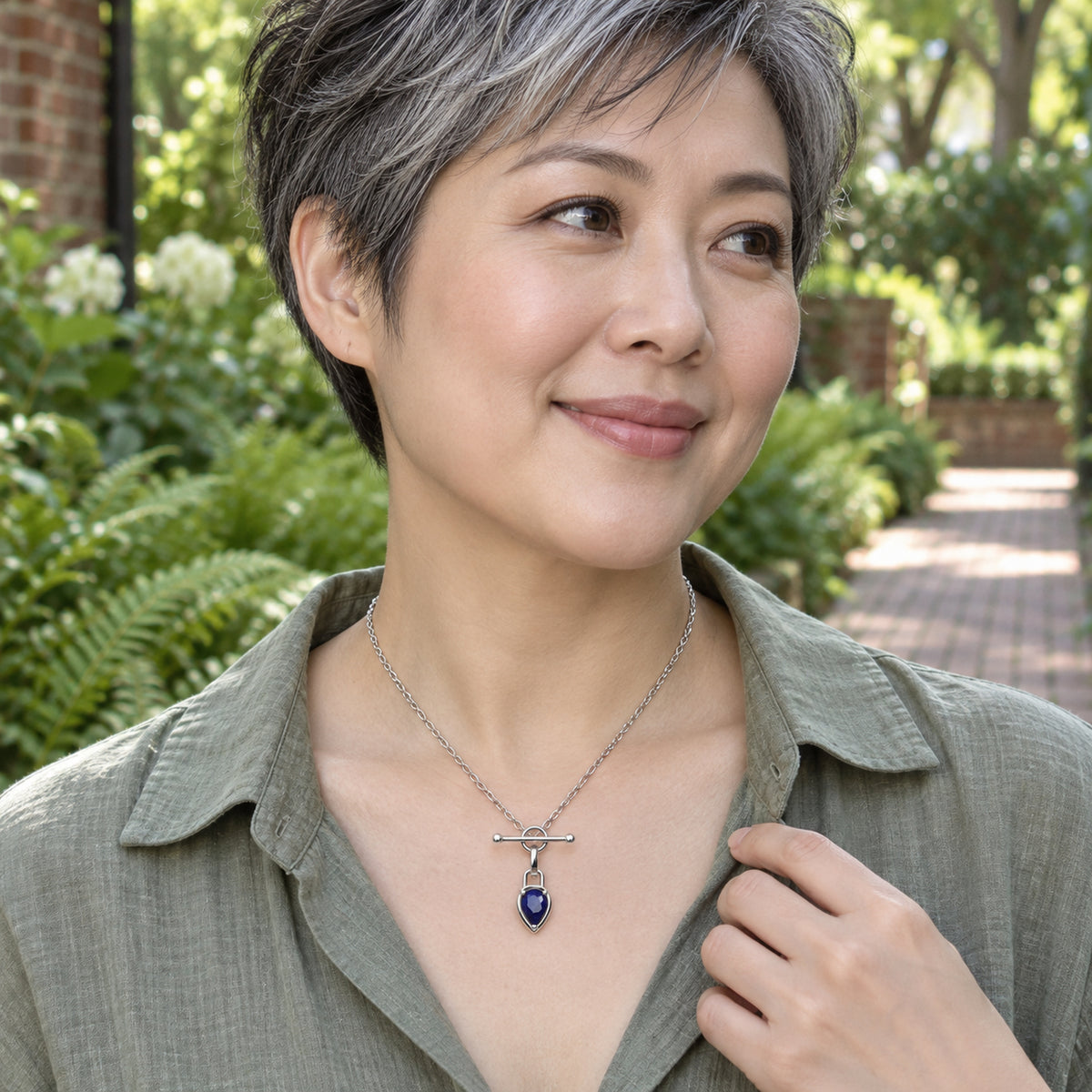 A woman in a sage linen blouse wears a silver-tone blue pendant necklace beside a leafy garden path.
