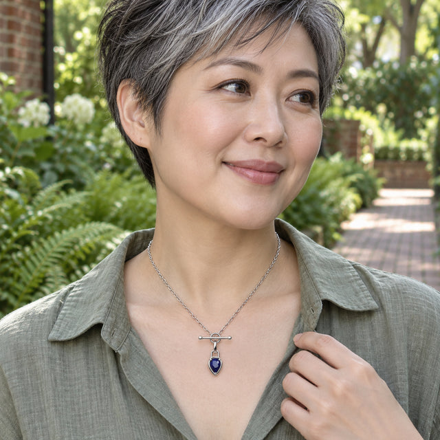 A woman in a sage linen blouse wears a silver-tone blue pendant necklace beside a leafy garden path.