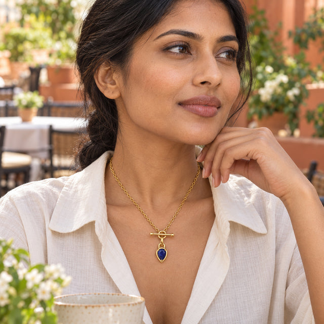 A woman in a cream blouse wears a gold-tone blue pendant necklace at an outdoor cafe table.