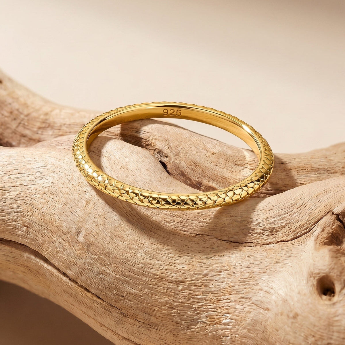 A yellow gold ring lies on a pale wooden surface with its patterned front edge facing the camera.