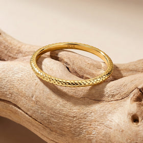 A yellow gold ring lies on a pale wooden surface with its patterned front edge facing the camera.