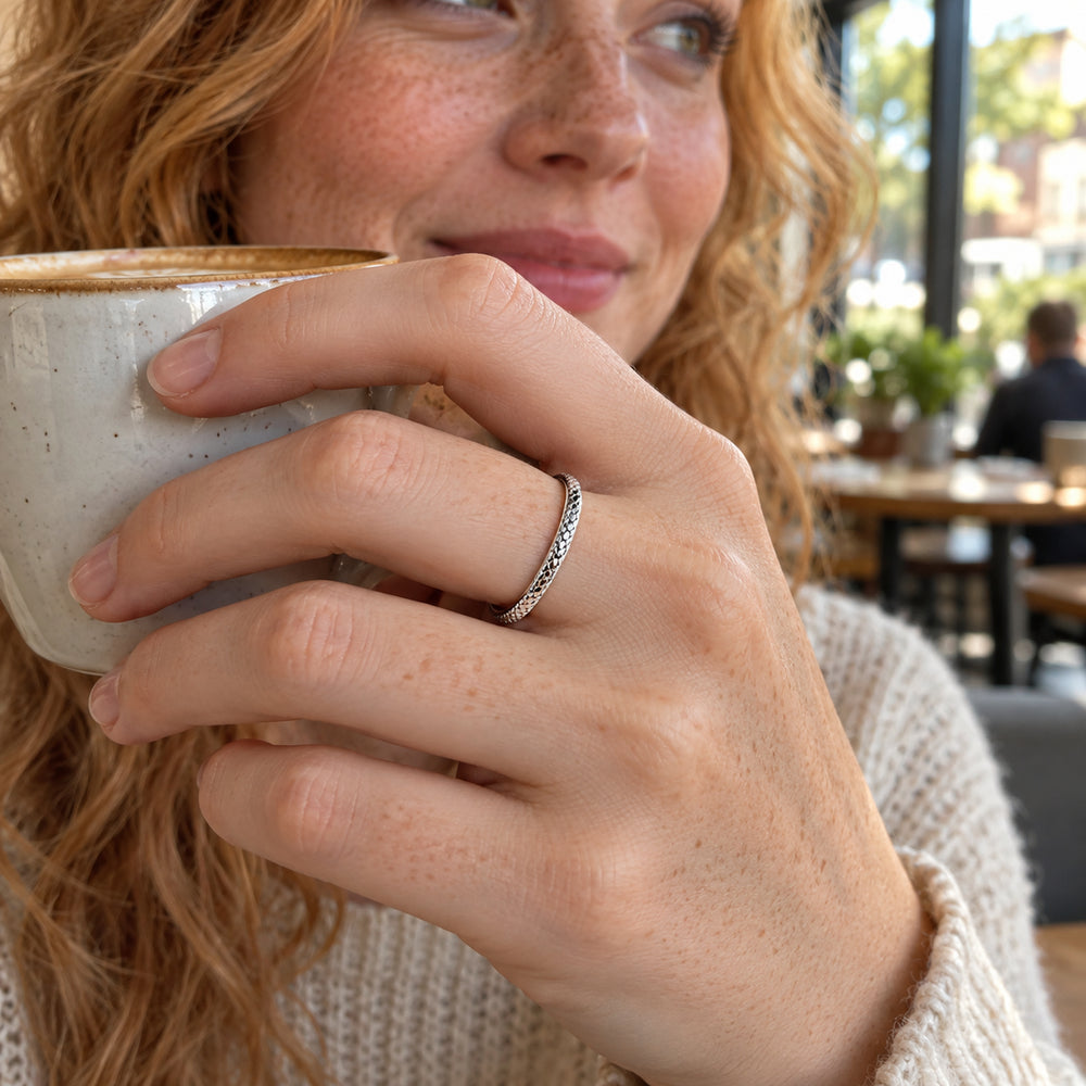 A woman with freckles wears a silver scale-textured ring while holding a ceramic cup by a cafe window.