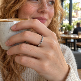 A woman with freckles wears a silver scale-textured ring while holding a ceramic cup by a cafe window.