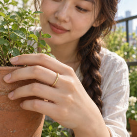 A yellow gold scale-textured ring is worn on a hand holding a terracotta plant pot on a leafy balcony.