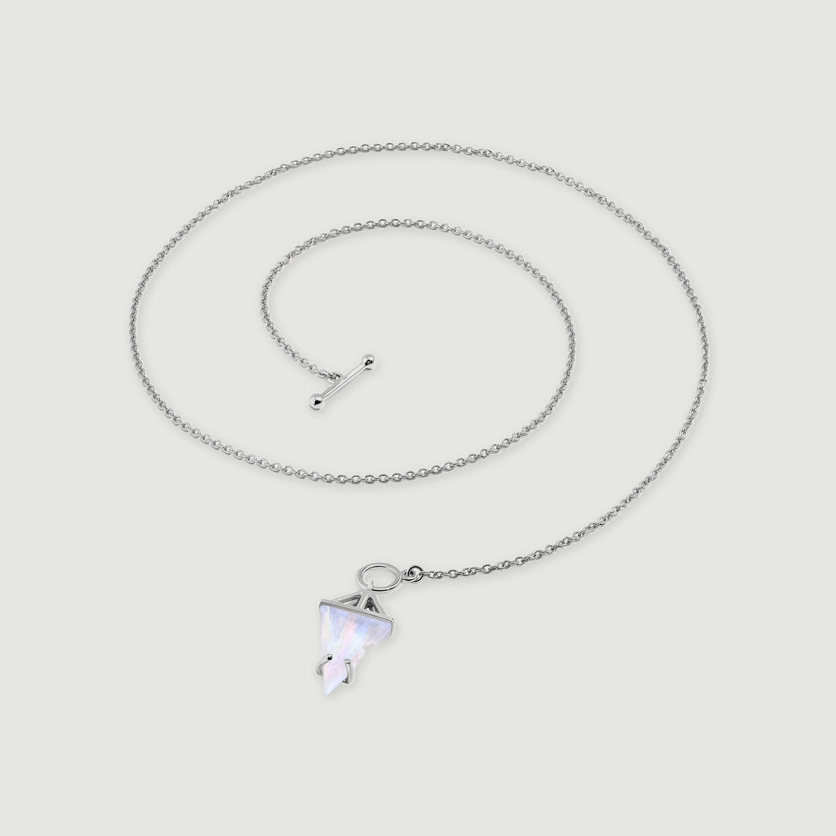 A sterling silver necklace lies in a loose coil with a triangular moonstone pendant and a polished toggle clasp on pale gray.