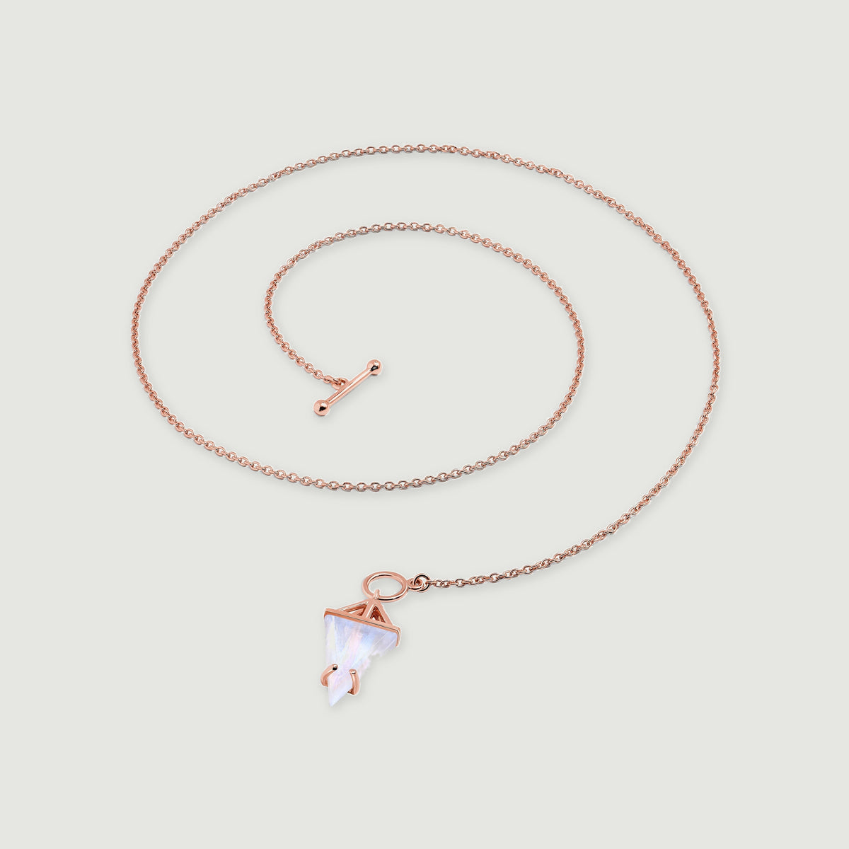 A rose gold vermeil necklace rests in an open coil, showing its linked chain and pale triangular moonstone on gray.