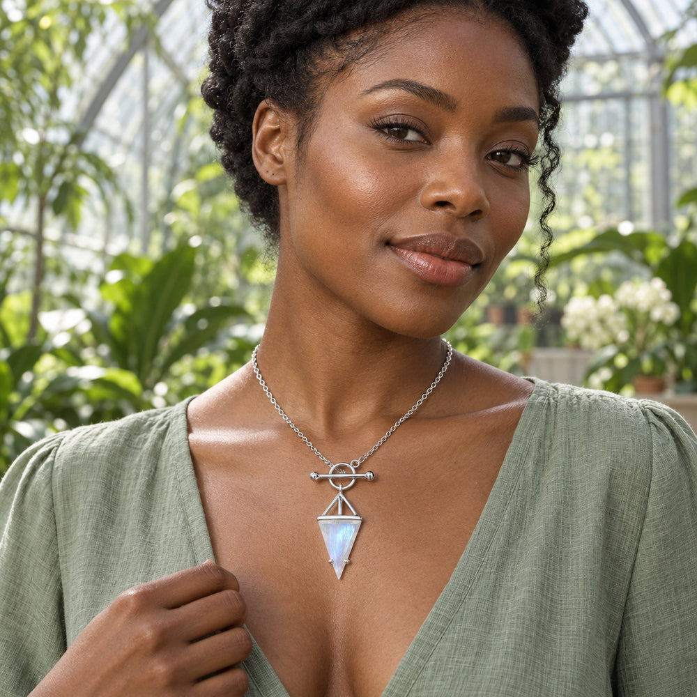A woman in a sage blouse wears a sterling silver necklace with a triangular moonstone pendant inside a leafy conservatory.
