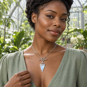 A woman in a sage blouse wears a sterling silver necklace with a triangular moonstone pendant inside a leafy conservatory.
