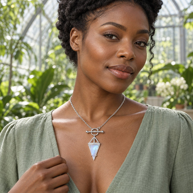A woman in a sage blouse wears a sterling silver necklace with a triangular moonstone pendant inside a leafy conservatory.